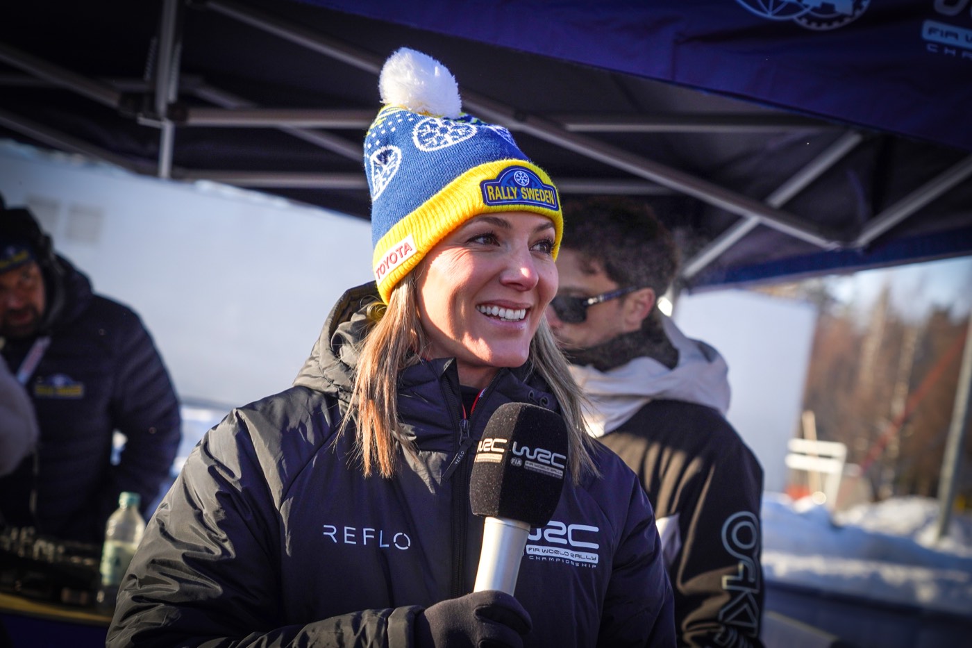 Presenter in a navy Reflo jacket and team bobble hat holding a microphone in the rally service park