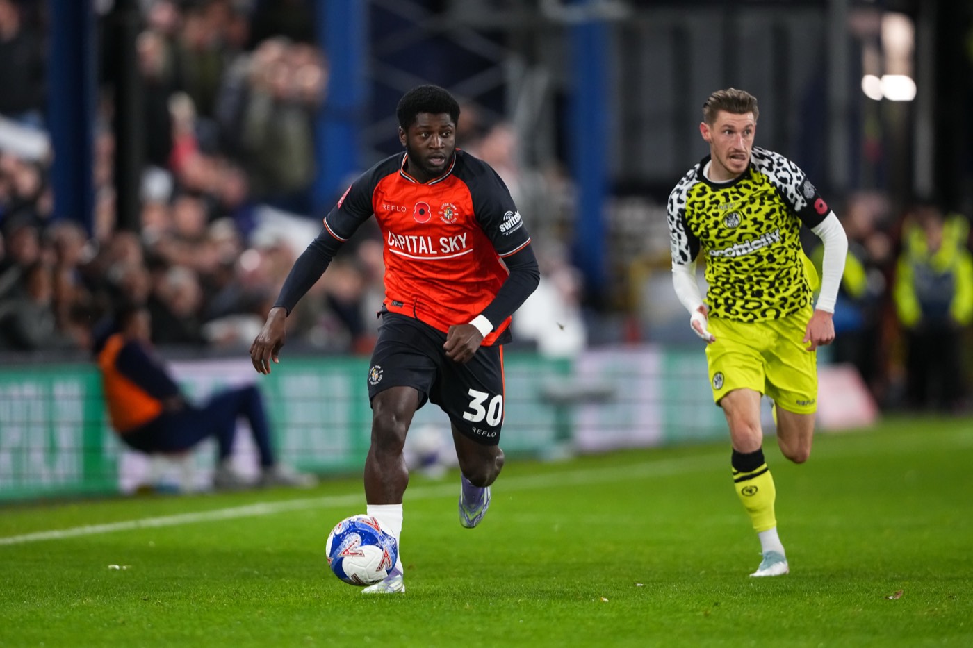 Two players competing for the ball on a floodlit pitch, one in the orange home shirt and one in the lime leopard-print away kit