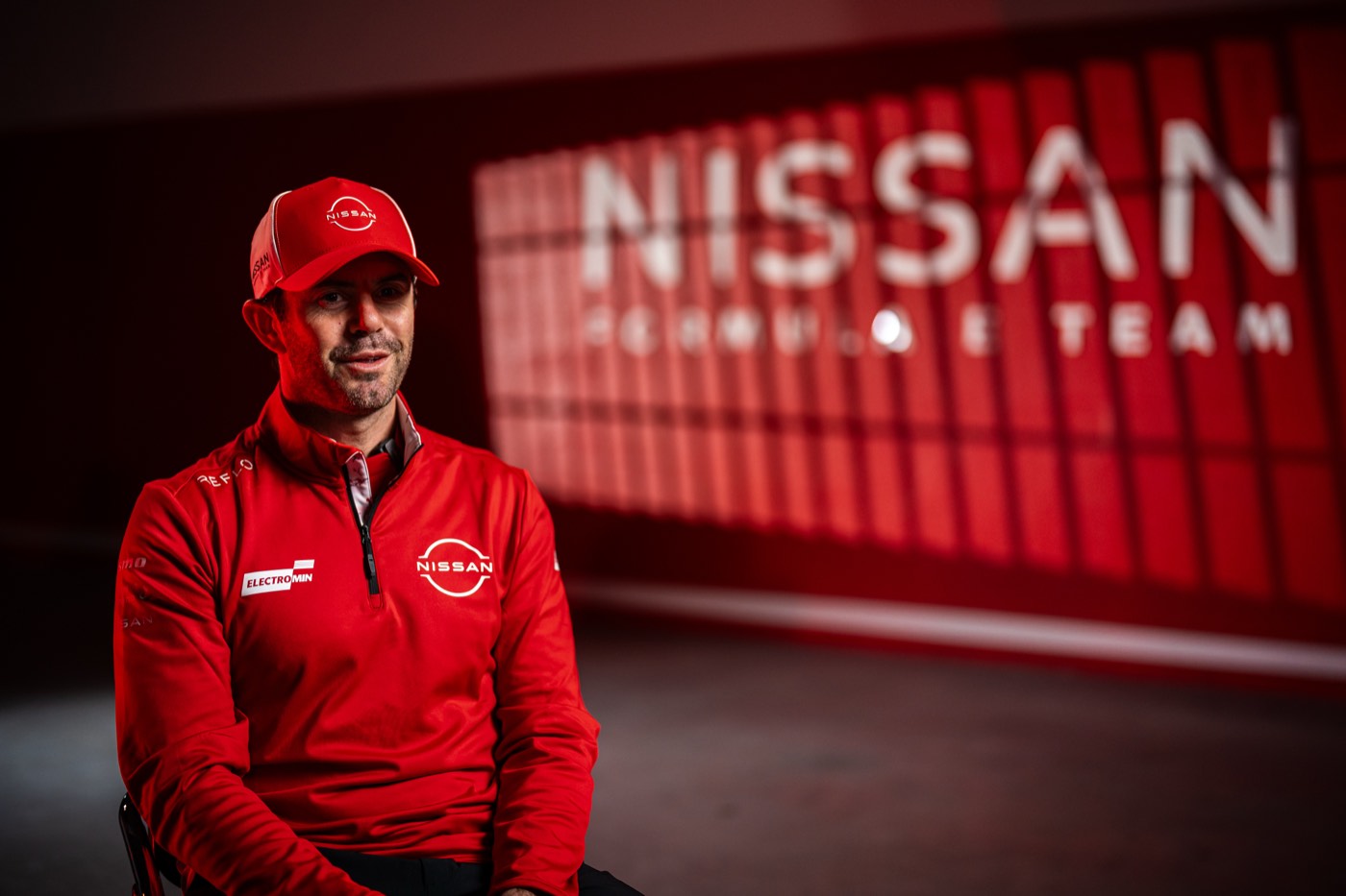 Team member in a red Reflo team jacket and cap in front of an illuminated Nissan Formula E Team sign