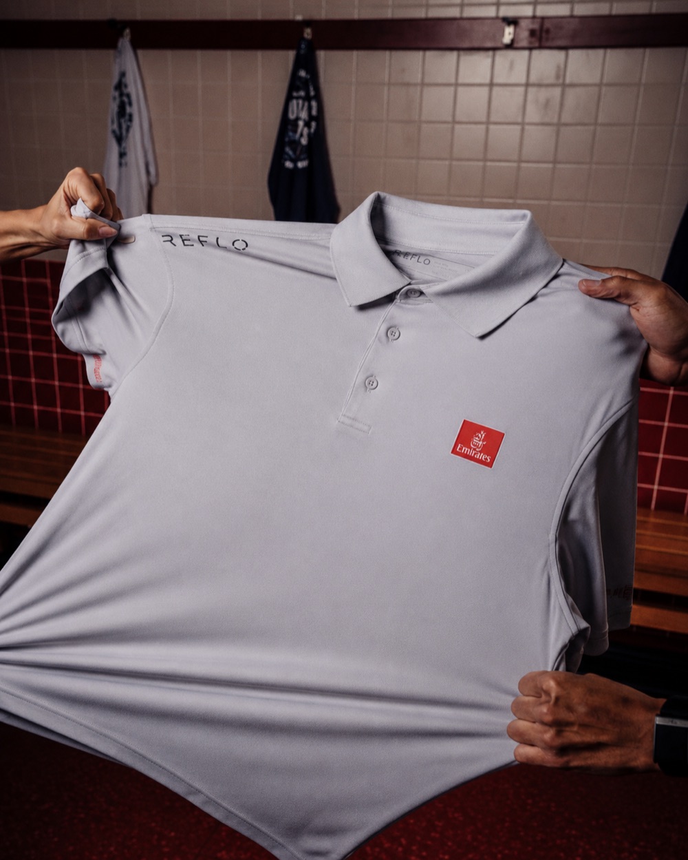 Grey Reflo polo shirt co-branded with the Emirates logo, held up in a locker room